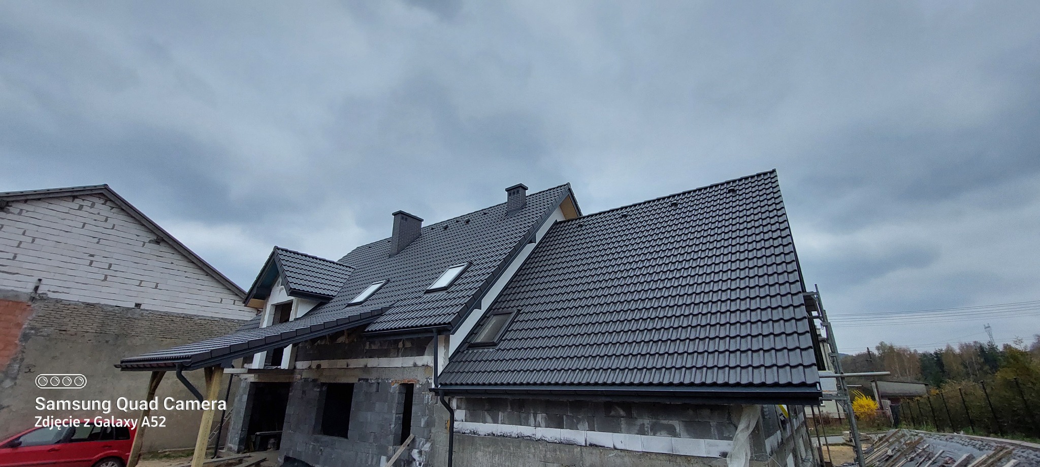 Nowy, grafitowy dach z dachówki ceramicznej na domu w trakcie budowy, widoczne kominy i okna dachowe, pochmurne niebo w tle. Ujęcie z dołu.