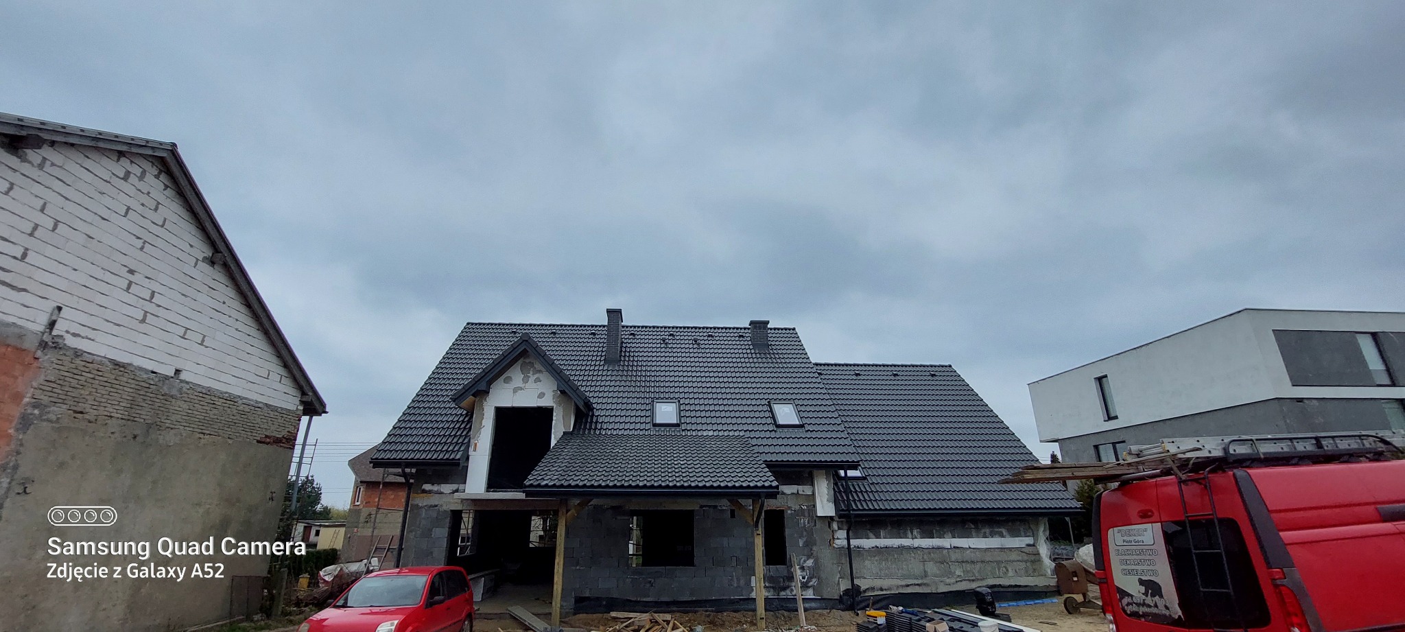 Dom w trakcie budowy z ciemnym dachem z dachówki, widoczne okna dachowe i komin. Częściowo otynkowana elewacja, czerwony samochód i budowlany bus na pierwszym planie.