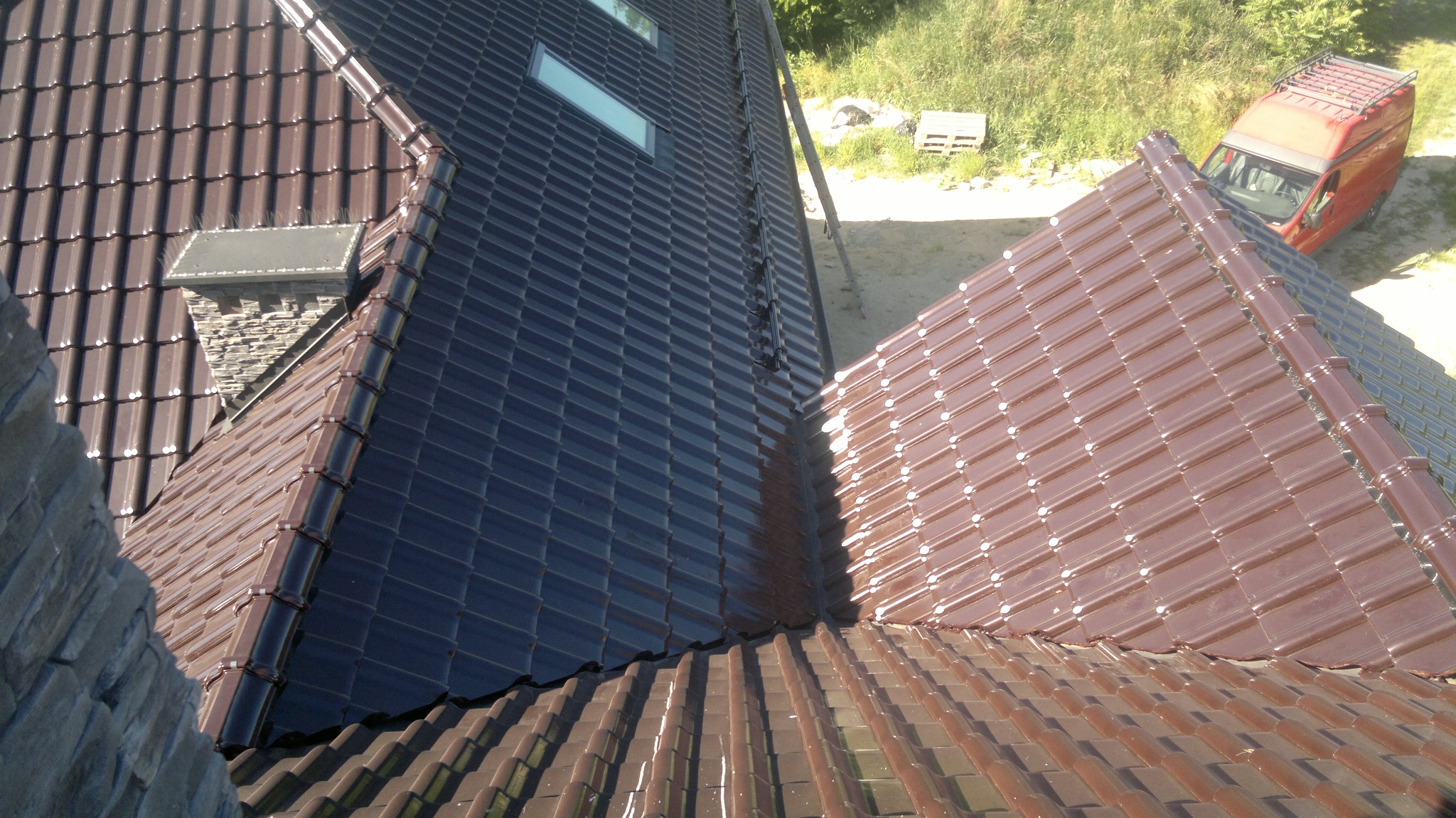 Nowy, brązowy dach z dachówki ceramicznej, z widocznym kominem i oknami dachowymi. Na drugim planie zielona trawa i czerwony samochód dostawczy.