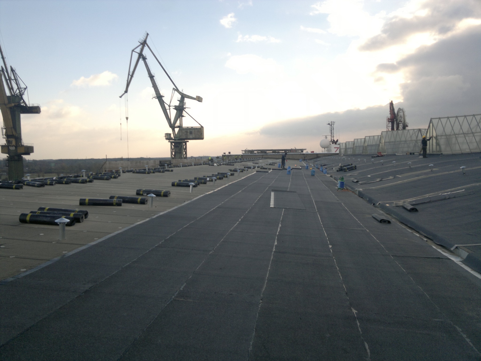 Rozległy, płaski dach pokryty papą termozgrzewalną w trakcie prac. W tle dźwigi portowe i pochmurne niebo. Widoczne rolki papy i pracownicy.
