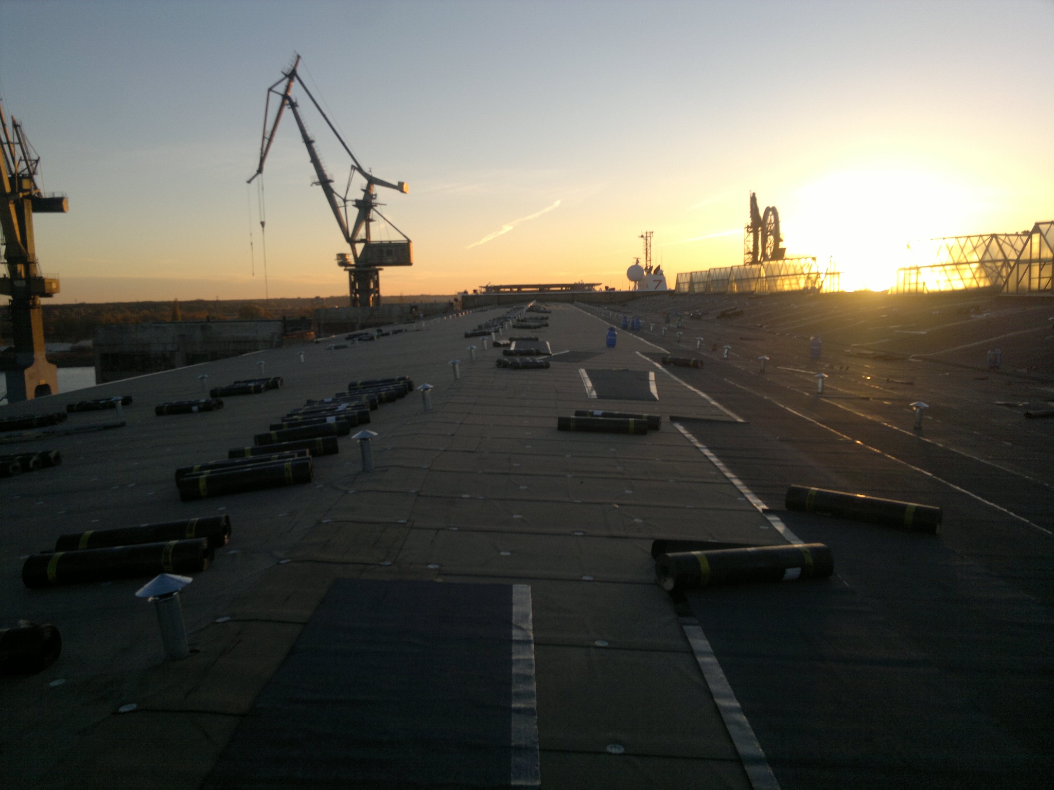 Rozległy dach przemysłowy w trakcie hydroizolacji, z rolkami papy bitumicznej i kominkami wentylacyjnymi, na tle zachodzącego słońca i dźwigów portowych.