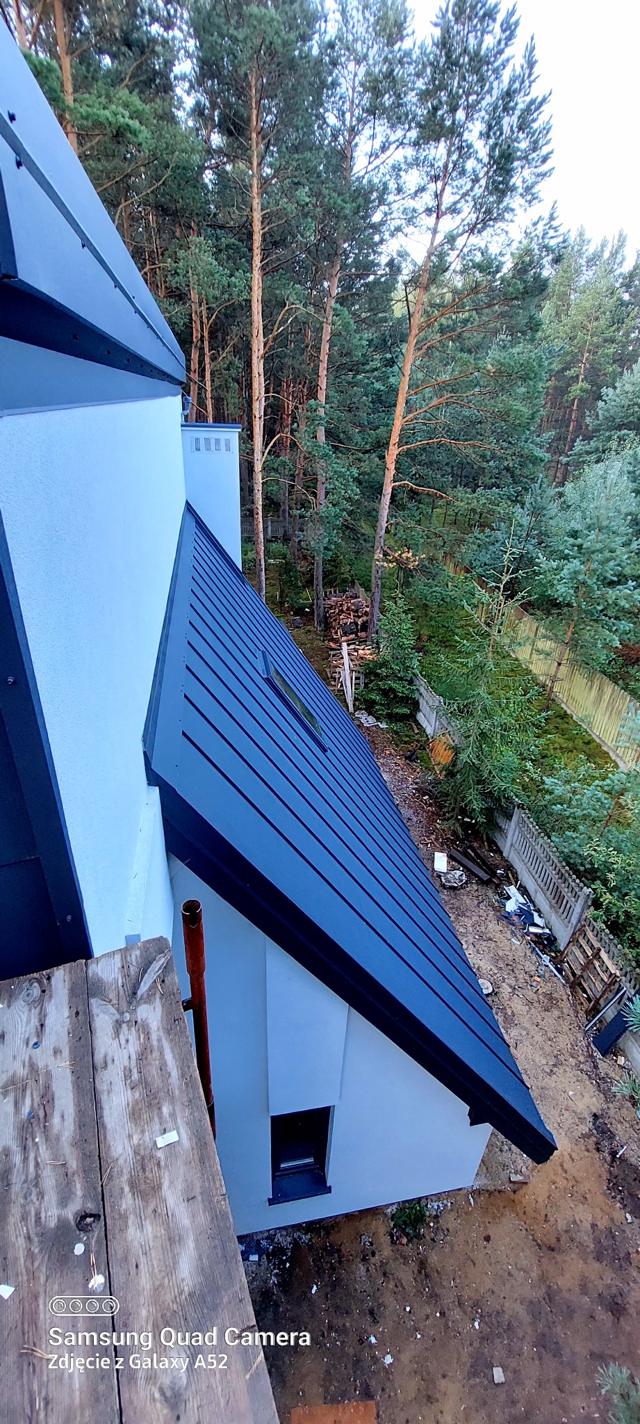 Nowoczesny dom z ciemnym, metalowym dachem widziany z góry. Widoczne drzewa w tle i fragment rusztowania. Dach z oknem dachowym.