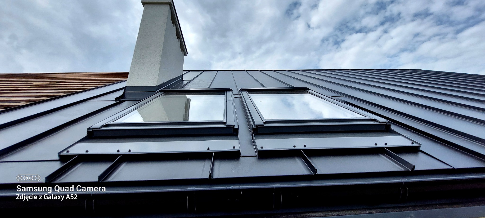 Czarny dach z blachy trapezowej z dwoma oknami dachowymi, widok z dołu na tle pochmurnego nieba i komina. Fragment dachu z drewnianą konstrukcją w tle.