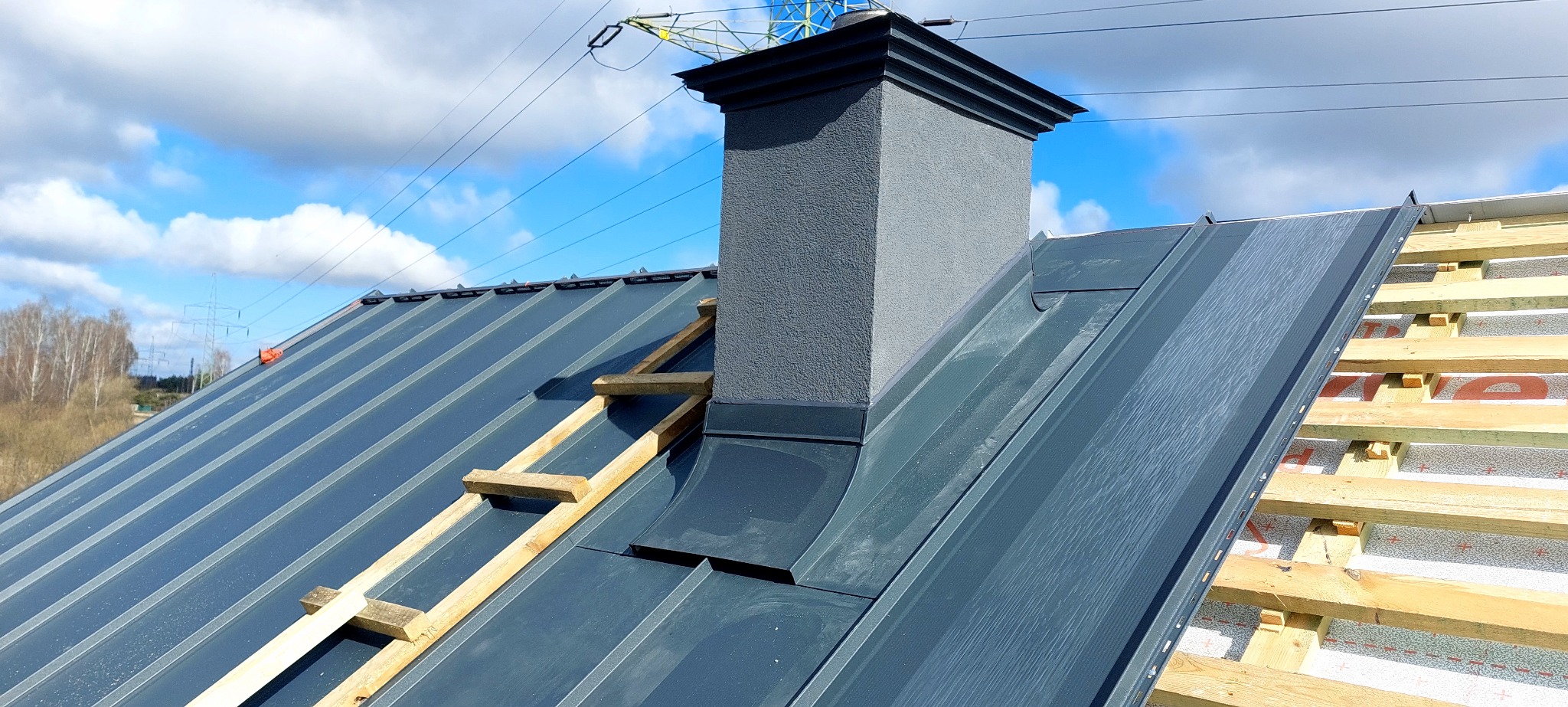 Szary dach z blachy trapezowej z kominem i drewnianą konstrukcją, widok z góry, błękitne niebo w tle. Detal pokrycia dachowego w trakcie montażu.