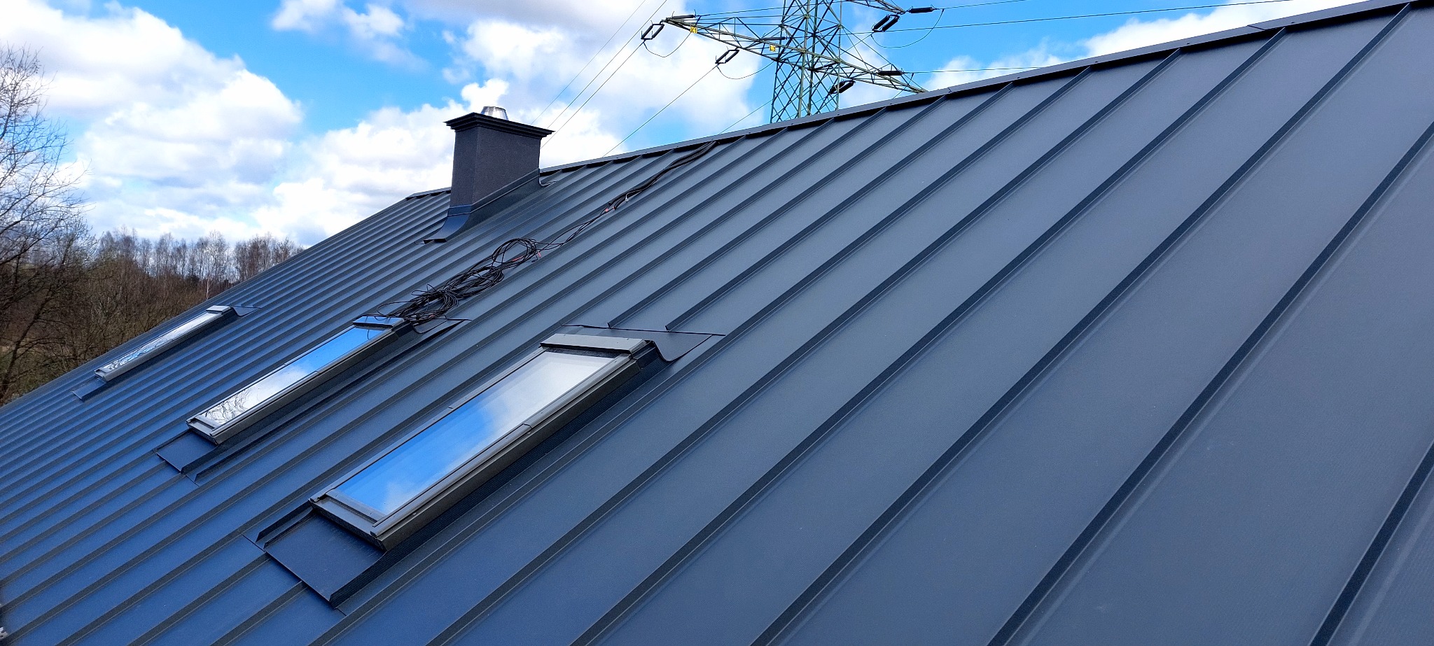 Szary dach z blachy trapezowej z trzema oknami dachowymi i kominem na tle błękitnego nieba z chmurami i słupem wysokiego napięcia w oddali.