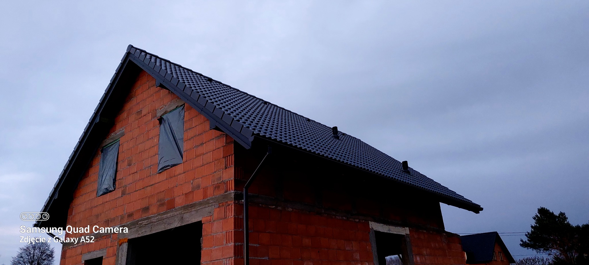 Ciemny dach z blachy na surowym budynku z czerwonej cegły pod pochmurnym niebem. Okna zabezpieczone folią. Widok z dołu na budynek w trakcie budowy.