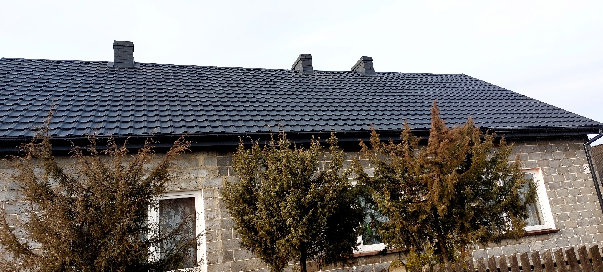 Dom z ciemnoszarym dachem z dachówki ceramicznej, widoczne kominy i rynny. Na pierwszym planie drzewa iglaste i fragment ogrodzenia. Ściany budynku z szarego pustaka.