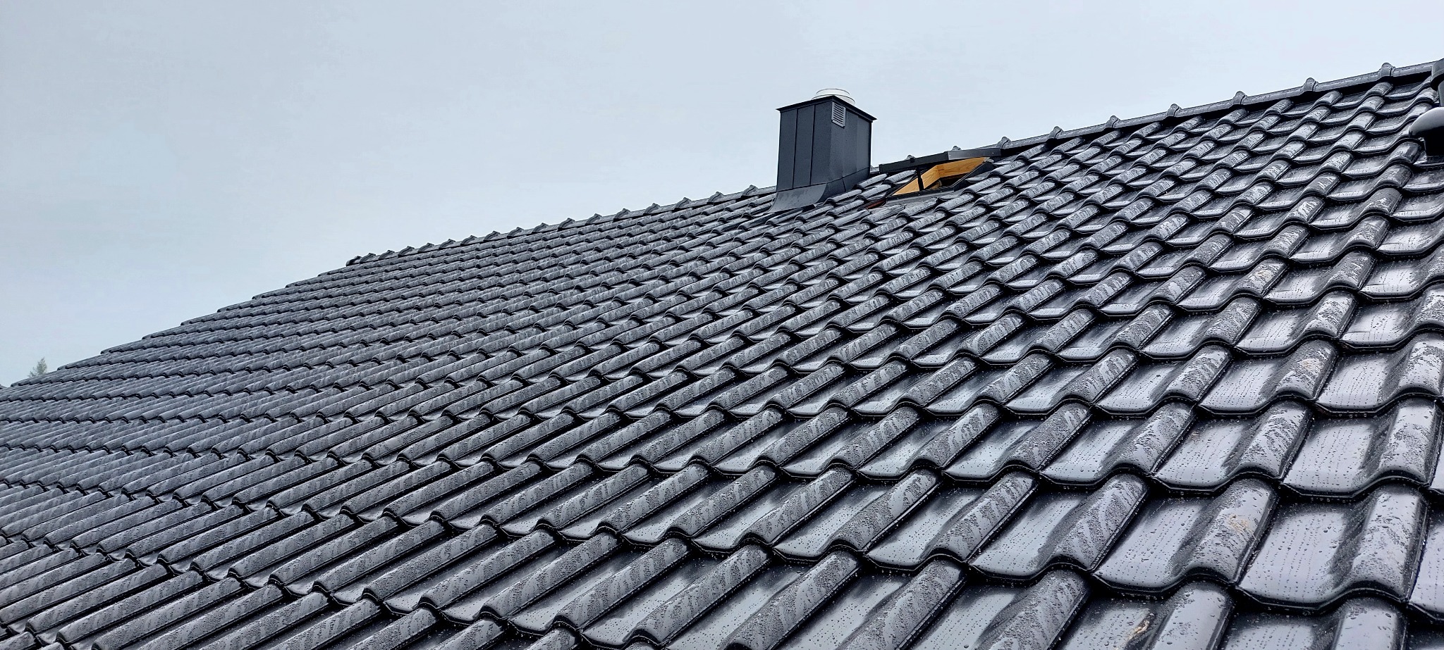 Czarny dach z dachówki ceramicznej, z widocznym kominem i fragmentem żółtej membrany dachowej. Dach lekko mokry po deszczu, pochmurne niebo w tle.