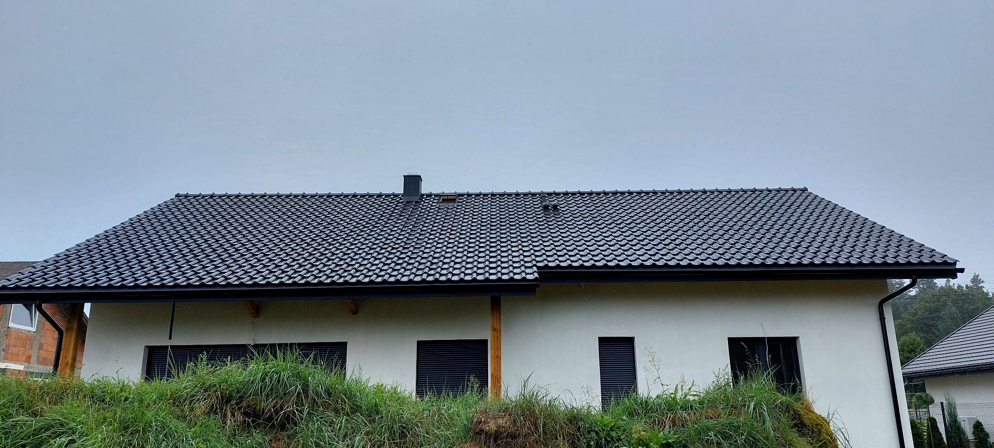 Dom jednorodzinny z ciemną dachówką ceramiczną, widok z dołu na dach i fragment elewacji. Na dachu komin i wywietrznik. W tle pochmurne niebo.