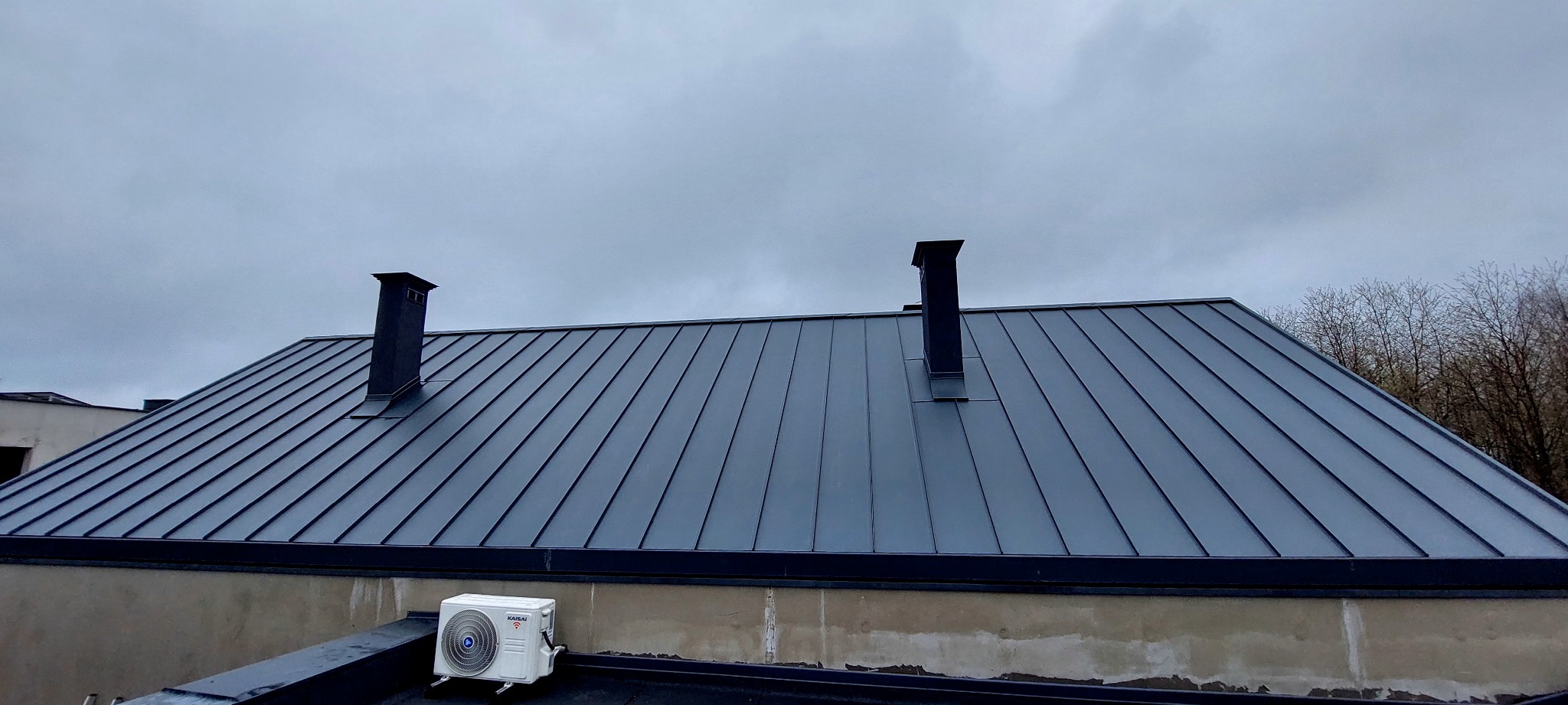 Nowoczesny dach z blachy na budynku z dwoma kominami. Widoczny klimatyzator na dachu. Pochmurne niebo w tle. Minimalistyczny design.