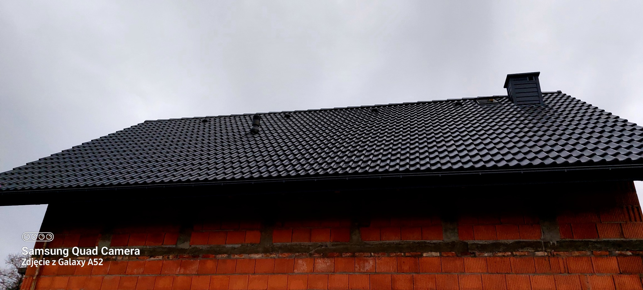 Nowy, czarny dach z dachówki ceramicznej na budynku w trakcie budowy, widoczny komin i rynna. Pochmurne niebo w tle. Ujęcie z dołu, podkreślające strukturę pokrycia.