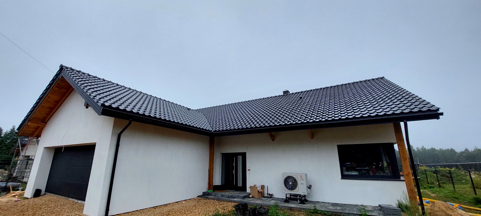 Dom jednorodzinny z ciemną dachówką karpiówką i czarnymi rynnami. Widoczny fragment elewacji, okno i drzwi wejściowe. Tło stanowi zieleń drzew i krzewów.