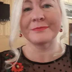 Portret kobiety z Ełku, blond włosy, czerwona szminka, czarna bluzka w kropki z kwiatowym haftem, czerwone kolczyki, w tle ściana z tapetą.