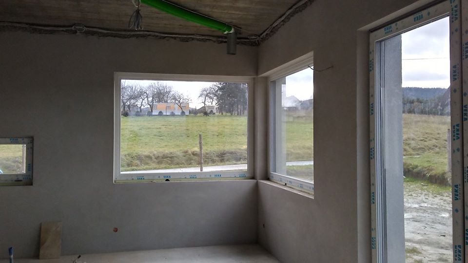 Wnętrze surowego domu po nałożeniu szarego tynku maszynowego na ściany, widoczne okna z widokiem na krajobraz za oknem, rury pod sufitem.