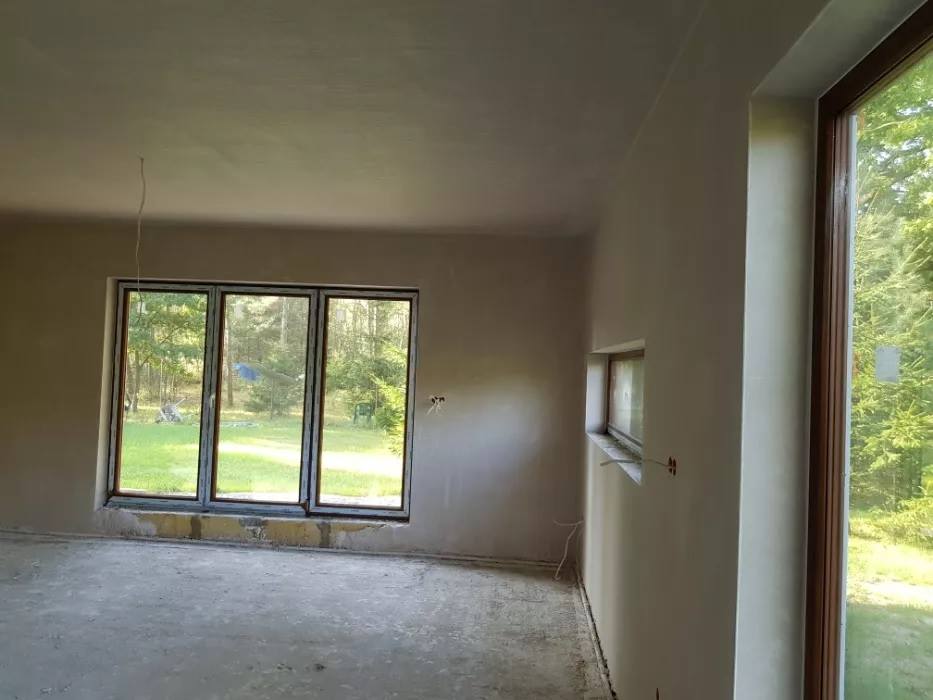 Wnętrze domu po nałożeniu gładzi gipsowej, widoczne okna z widokiem na zieleń, surowy beton na podłodze i wystające przewody elektryczne.