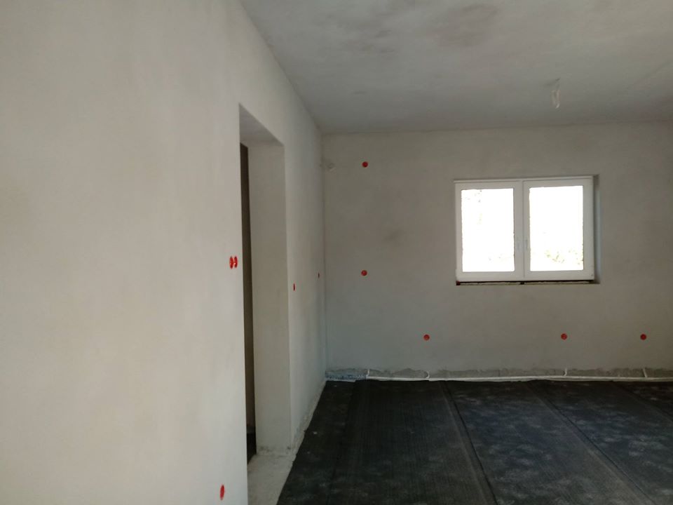 Wnętrze pomieszczenia po nałożeniu tynków maszynowych, widoczne gładkie, jasne ściany z czerwonymi punktami odniesienia, czarna folia na podłodze, okno w tle.