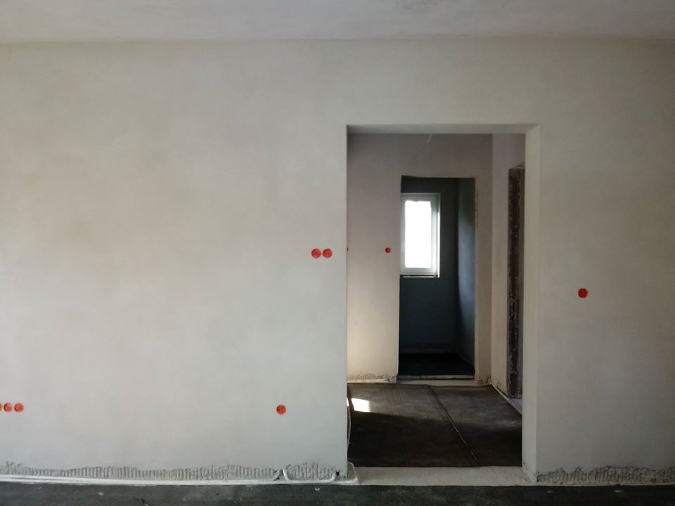 Świeżo położony gładki tynk na ścianie wewnątrz budynku, widoczne oznaczenia punktów elektrycznych w postaci czerwonych kropek, w tle widok na korytarz z oknem.