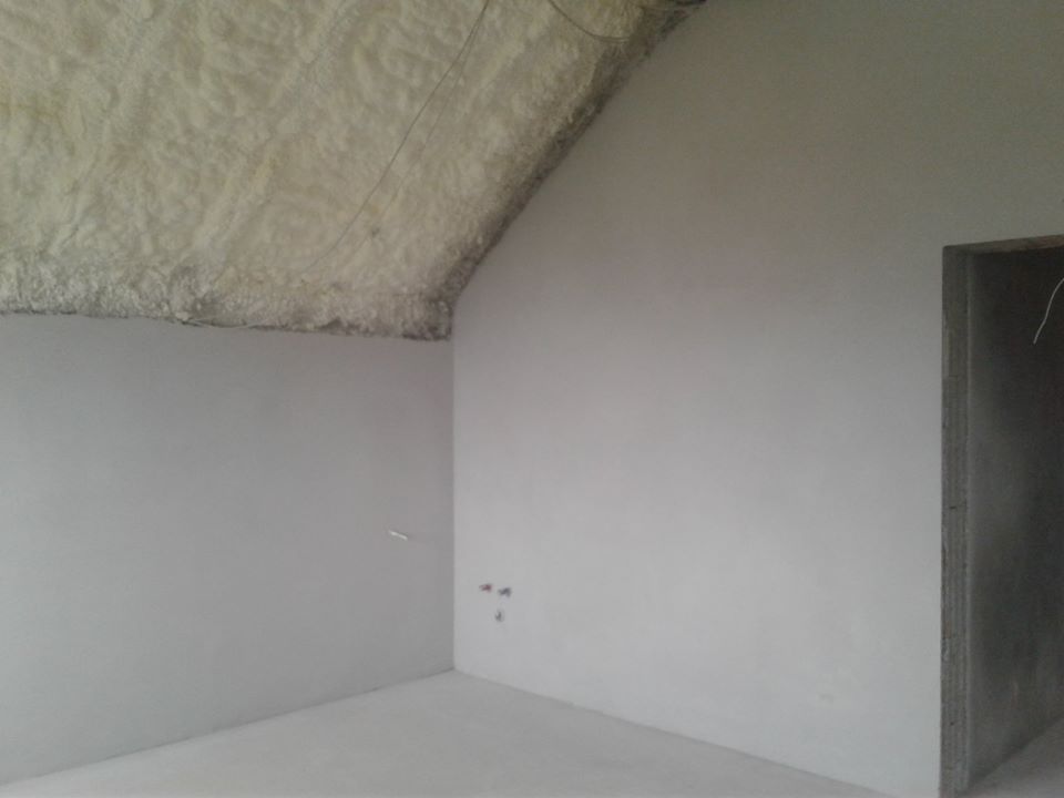 Pokój po nałożeniu izolacji piankowej na poddaszu i wstępnym wykończeniu ścian szarym tynkiem, widoczne niedoskonałości na suficie i ścianach.