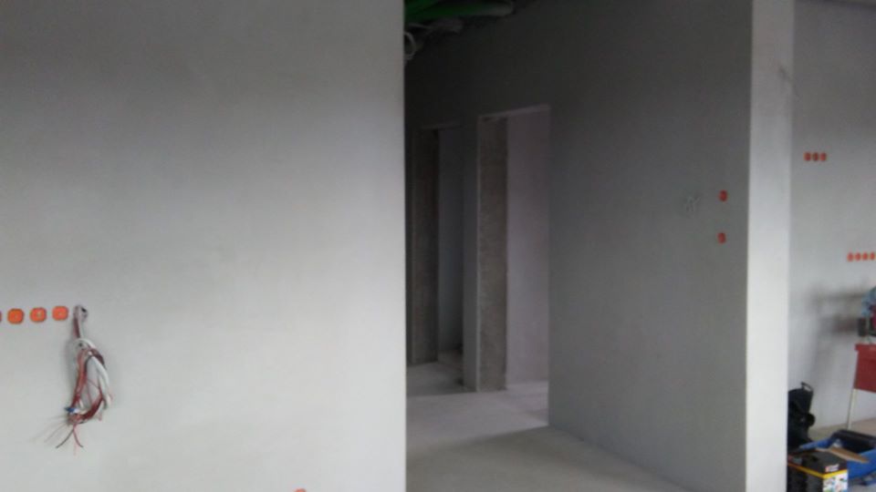 Świeżo otynkowane ściany w surowym wnętrzu, widoczne wystające przewody elektryczne i otwory pod gniazdka, w tle widoczne kolejne pomieszczenia w stanie deweloperskim.