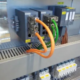 Panel sterowniczy z modułami Siemens SCALANCE i SIMATIC, połączony przewodami w kolorach pomarańczowym i zielonym oraz zabezpieczeniami z żółtymi etykietami.