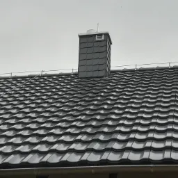 Szary dach z dachówki karpiówki i komin w tym samym kolorze. Widoczne zabezpieczenia przeciwśniegowe. Pochmurne niebo w tle.