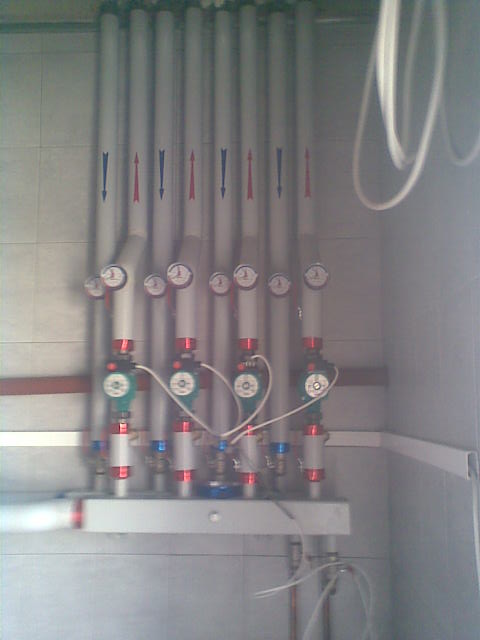 Rozdzielacz ogrzewania podłogowego z manometrami i pompami obiegowymi, widoczne rury z oznaczeniami przepływu w kolorze czerwonym i niebieskim, zamontowany przy szarej ścianie wyłożonej kafelkami.