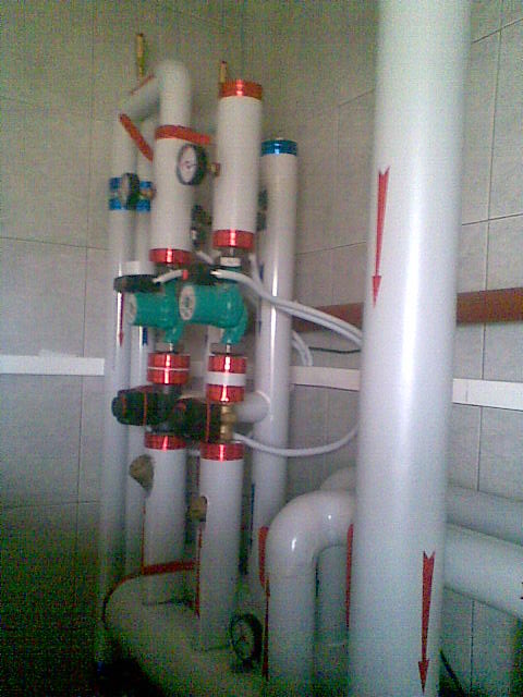 Złożona instalacja grzewcza z białych rur z czerwonymi i niebieskimi akcentami, zawierająca pompy obiegowe i manometry, umieszczona w pomieszczeniu z wykafelkowanymi ścianami.