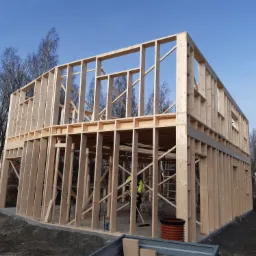 Wznoszenie drewnianej konstrukcji budynku w systemie szkieletowym, widoczny szkielet ścian i stropów, pracownik w odblaskowej kamizelce wewnątrz konstrukcji, błękitne niebo w tle.