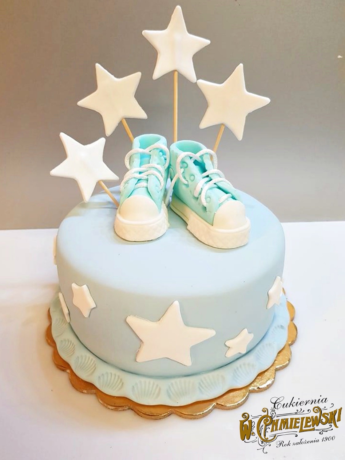 Tort w Lublinie z dekoracjami: niebieski tort z białymi gwiazdami i parą turkusowych butów z lukru na wierzchu, dodatkowo pięć białych gwiazd na patykach.
