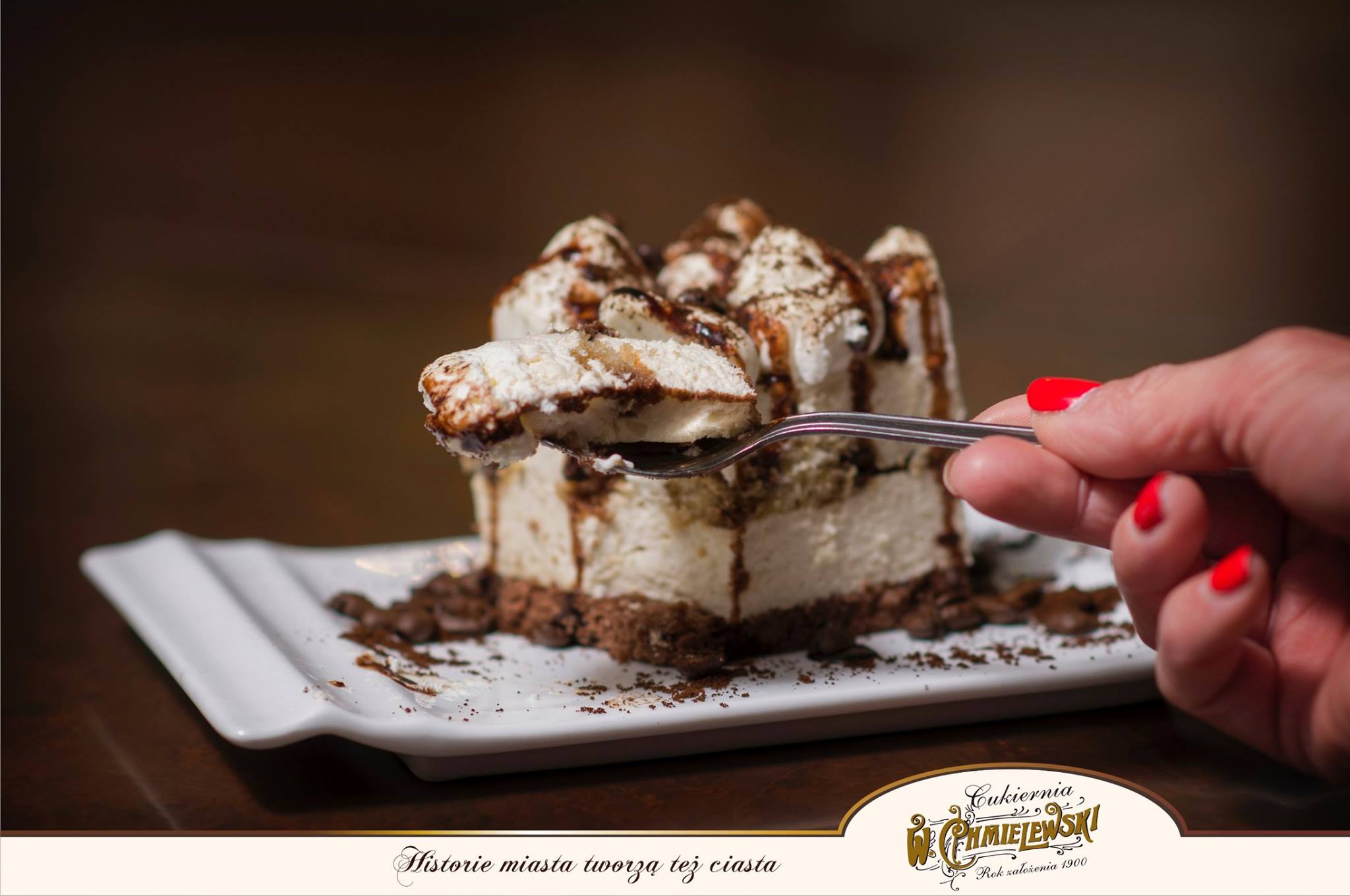 Kremowy deser z polewą czekoladową na białym talerzu, unoszony na łyżeczce przez dłoń z czerwonymi paznokciami, widoczne logo firmy i slogan reklamowy.