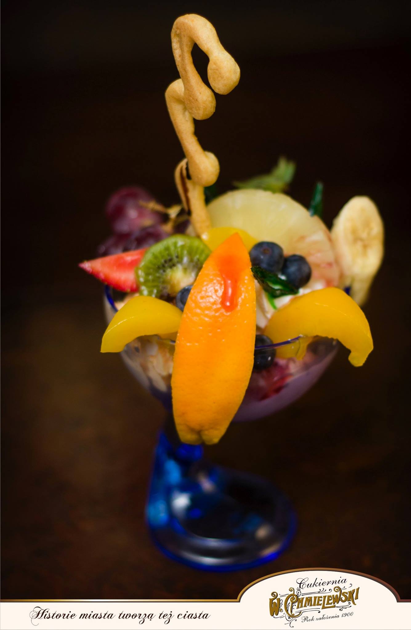 Deser lodowy w pucharku na nóżce, udekorowany owocami: kiwi, truskawką, winogronami, ananasem, bananem, brzoskwiniami w syropie i pomarańczą z sosem, zwieńczony kruchym ciastkiem w kształcie litery S.