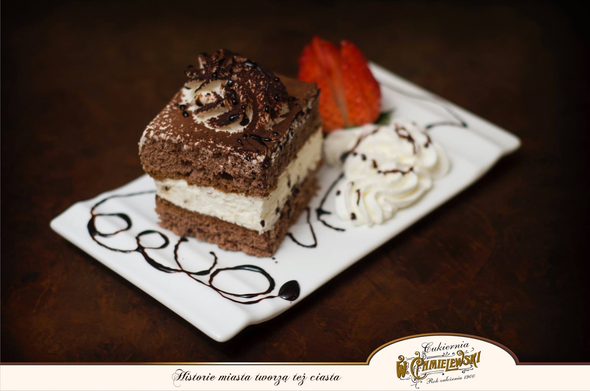 Kwadratowy kawałek ciasta czekoladowego z kremem, udekorowany bitą śmietaną, polewą czekoladową i truskawką na białym talerzu z dekoracyjnymi kleksami czekolady.