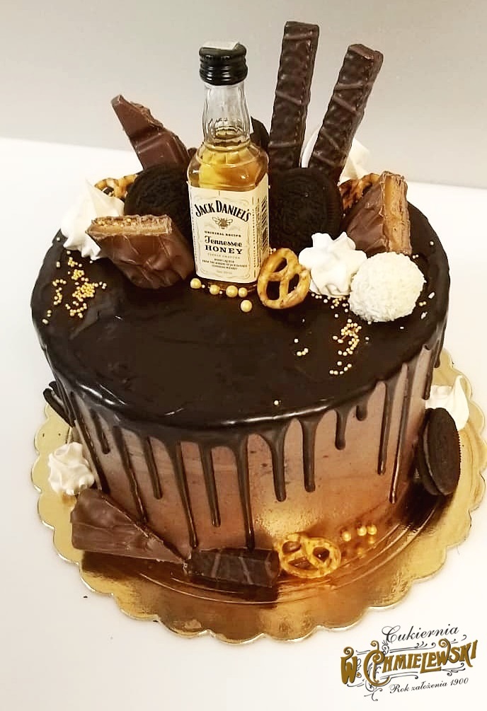 Tort czekoladowy z polewą, dekorowany miniaturową butelką Jack Daniel's Honey, batonikami, ciastkami Oreo, preclami i złotymi kuleczkami, umieszczony na złotej podkładce z widocznym logo cukierni.