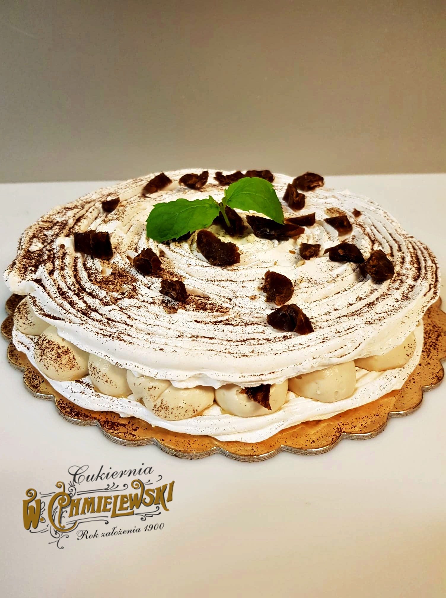 Okrągły tort bezowy z kremem, posypany kakao i kawałkami suszonych śliwek, udekorowany listkami mięty, na złotej podkładce. Widoczne logo 'Cukiernia W. Chmielewski. Rok założenia 1900'.