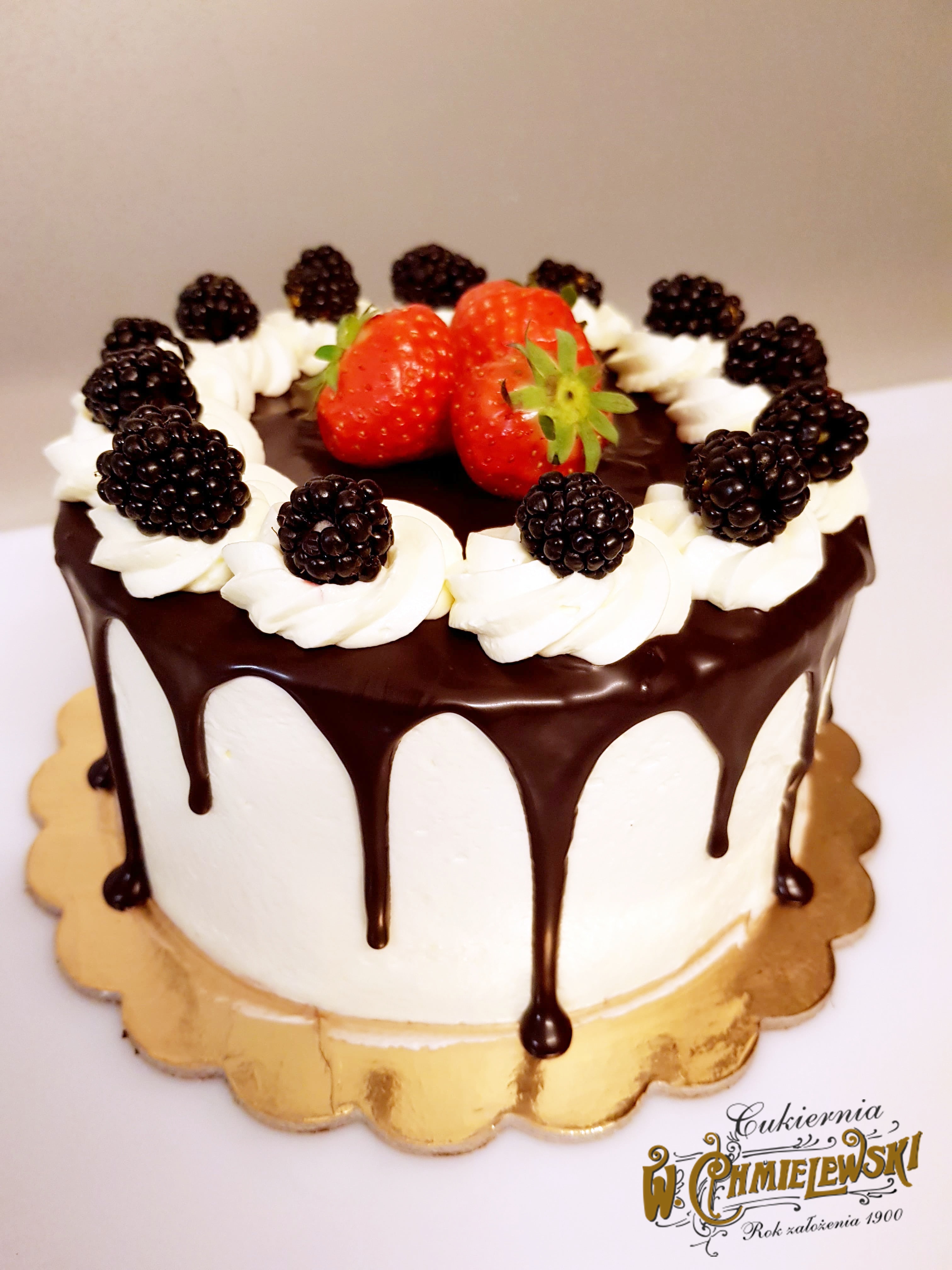 Tort z białym kremem, polewą czekoladową, jeżynami i truskawkami, umieszczony na złotej podkładce, widoczne logo firmy na podkładce