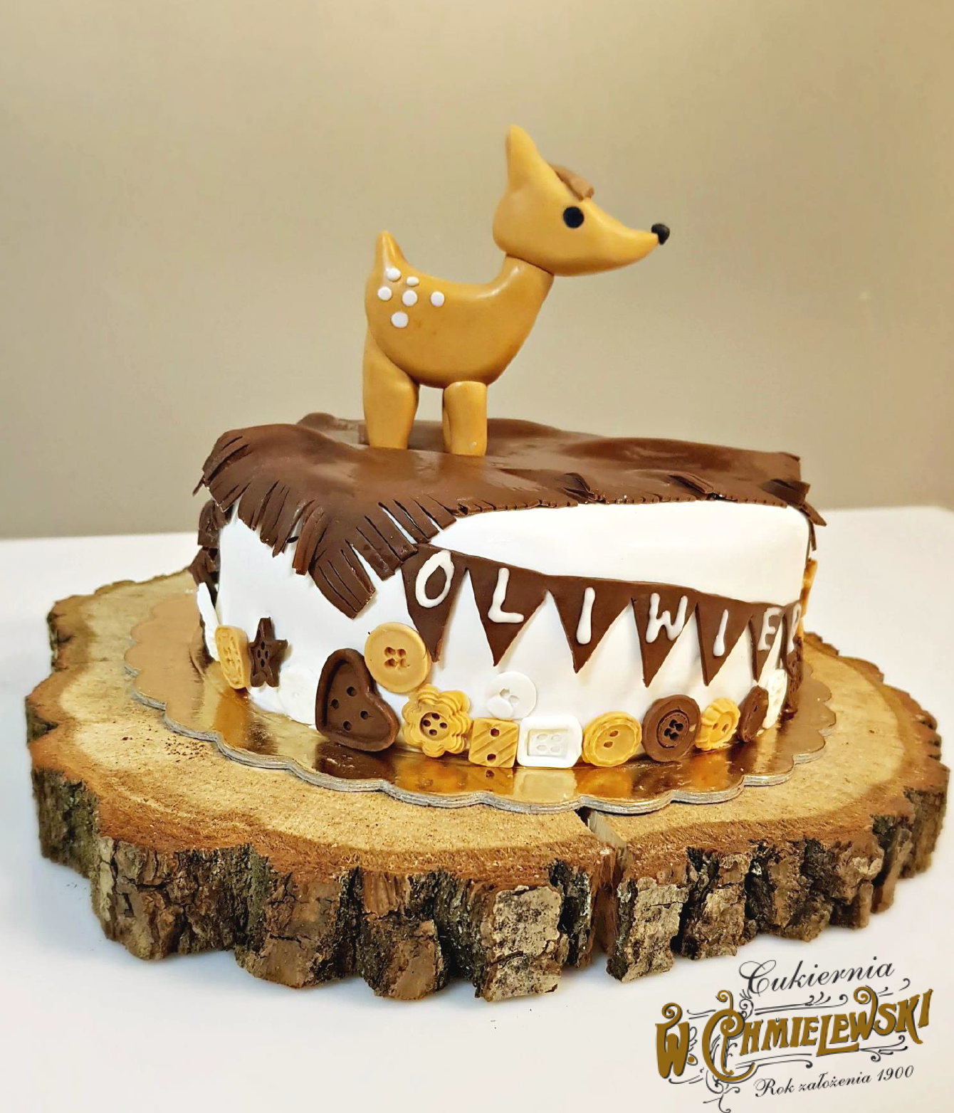 Tort urodzinowy z dekoracją w postaci brązowego jelenia z lukru, imieniem 'Oliwia' na białym tle, ozdobiony czekoladowymi frędzlami, guzikami i gwiazdkami, umieszczony na drewnianym plastrze.