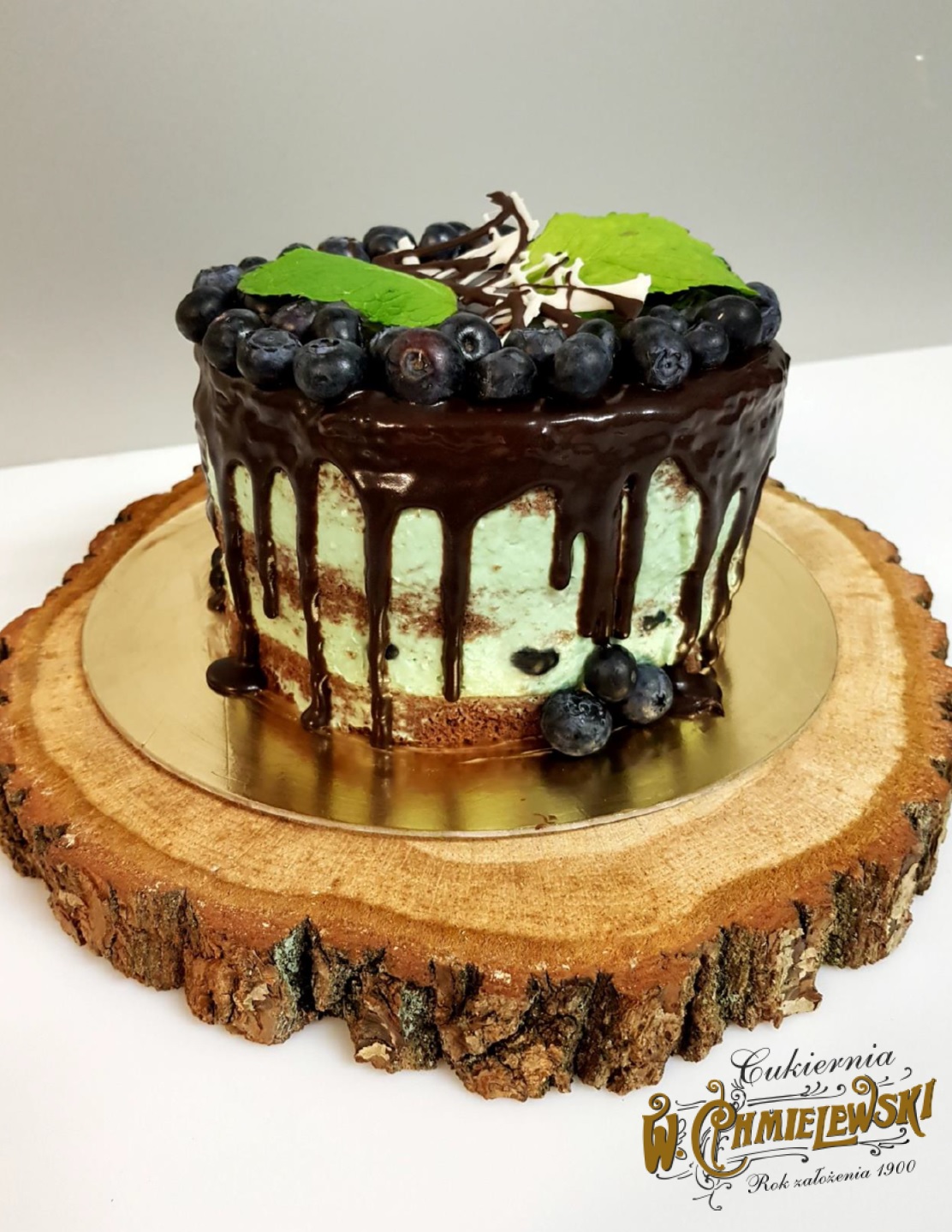 Mały tort z miętowym kremem, polany czekoladą i udekorowany borówkami oraz listkami mięty, umieszczony na drewnianym plastrze.
