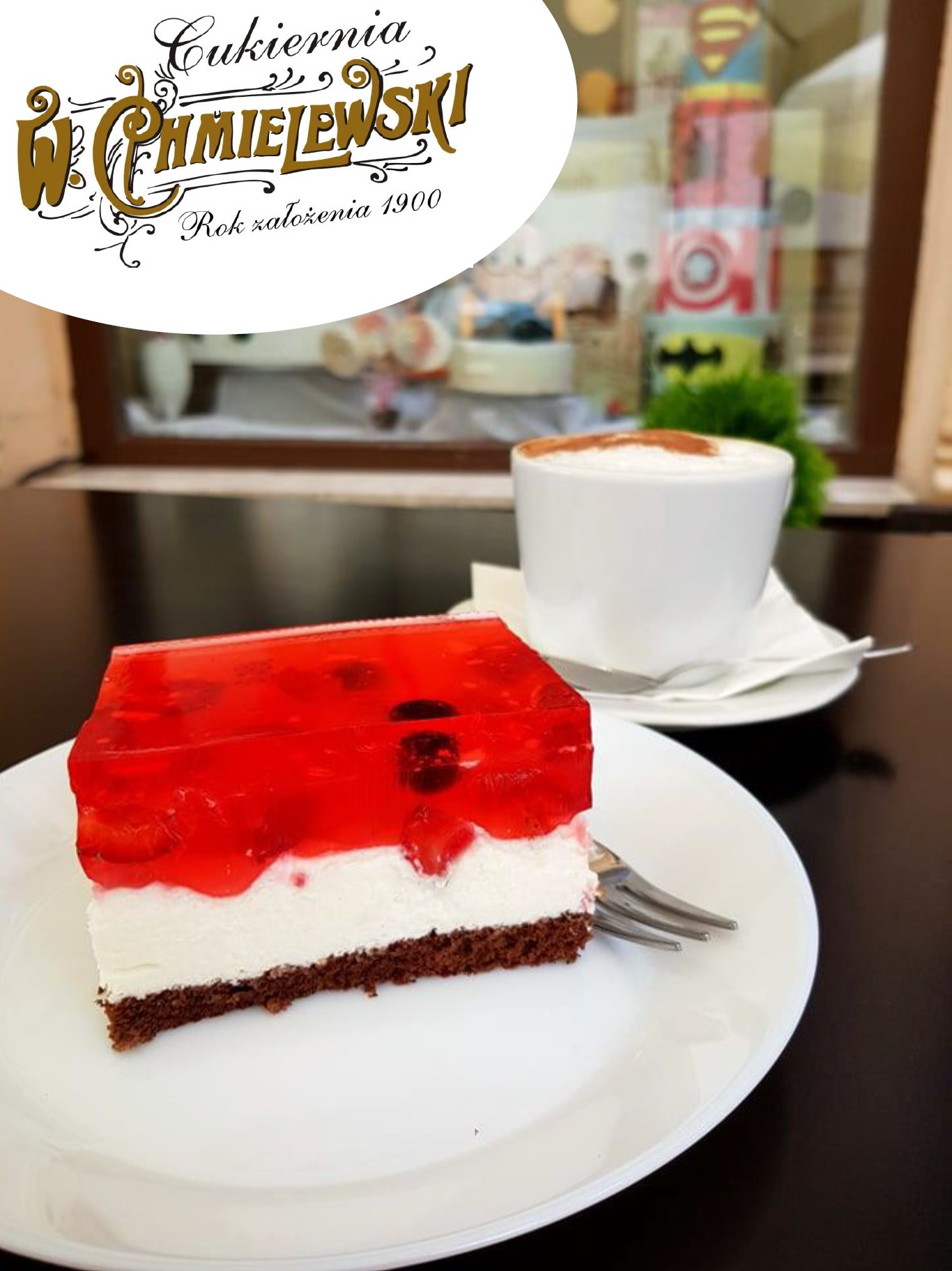 Kawałek ciasta z galaretką wiśniową na białym talerzu obok filiżanki cappuccino, w tle logo cukierni i dekoracje okienne.