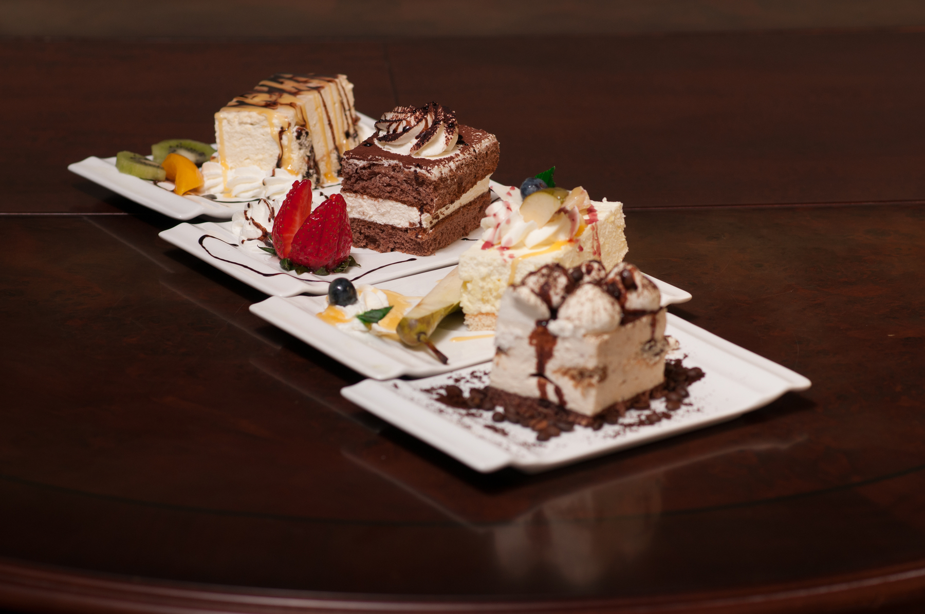 Cztery porcje różnorodnych ciast na białych, kwadratowych talerzykach, udekorowane owocami i polewami, ułożone na ciemnym, drewnianym stole.