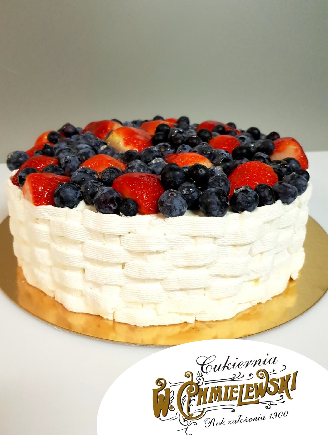 Tort z kremem imitującym plecionkę koszyka, udekorowany świeżymi truskawkami i borówkami na złotym podkładzie.
