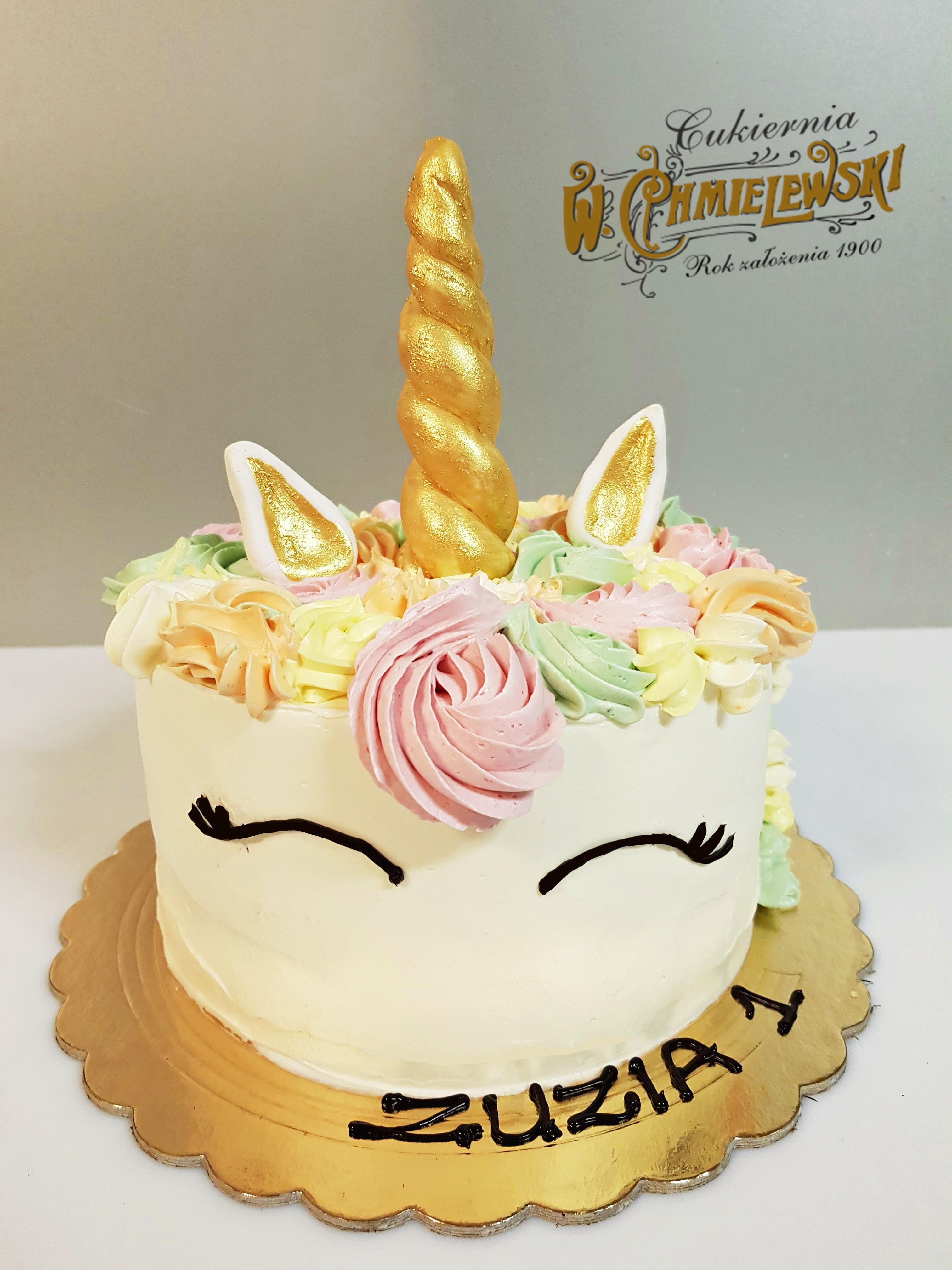 Tort jednorożec z kremowymi zdobieniami w pastelowych kolorach, złotym rogiem i uszami, na złotym podkładzie z napisem 'Zuzia 1', w tle widoczne logo firmy.