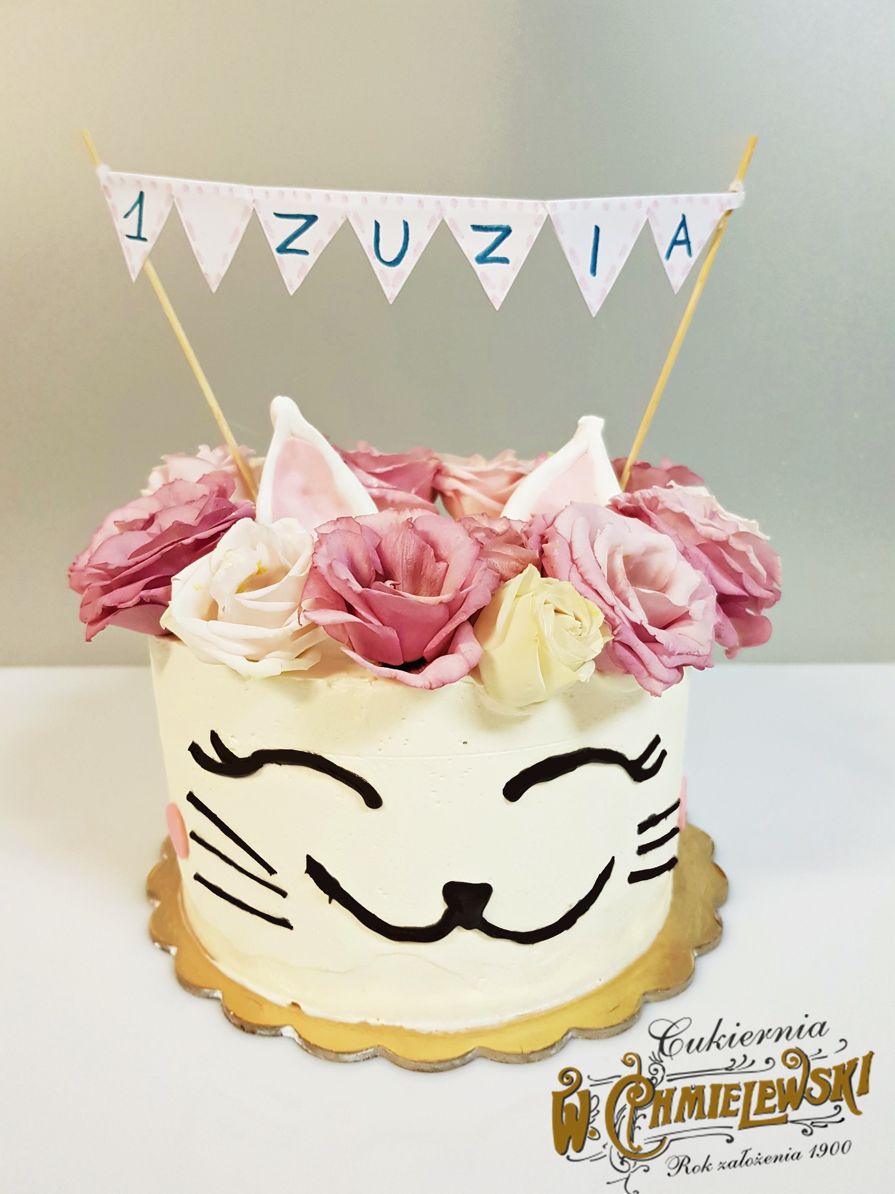 Tort urodzinowy dla rocznej Zuzi w kształcie kota, ozdobiony różowymi różami i uszami z masy cukrowej, na złotym podkładzie, z widocznym logo firmy.