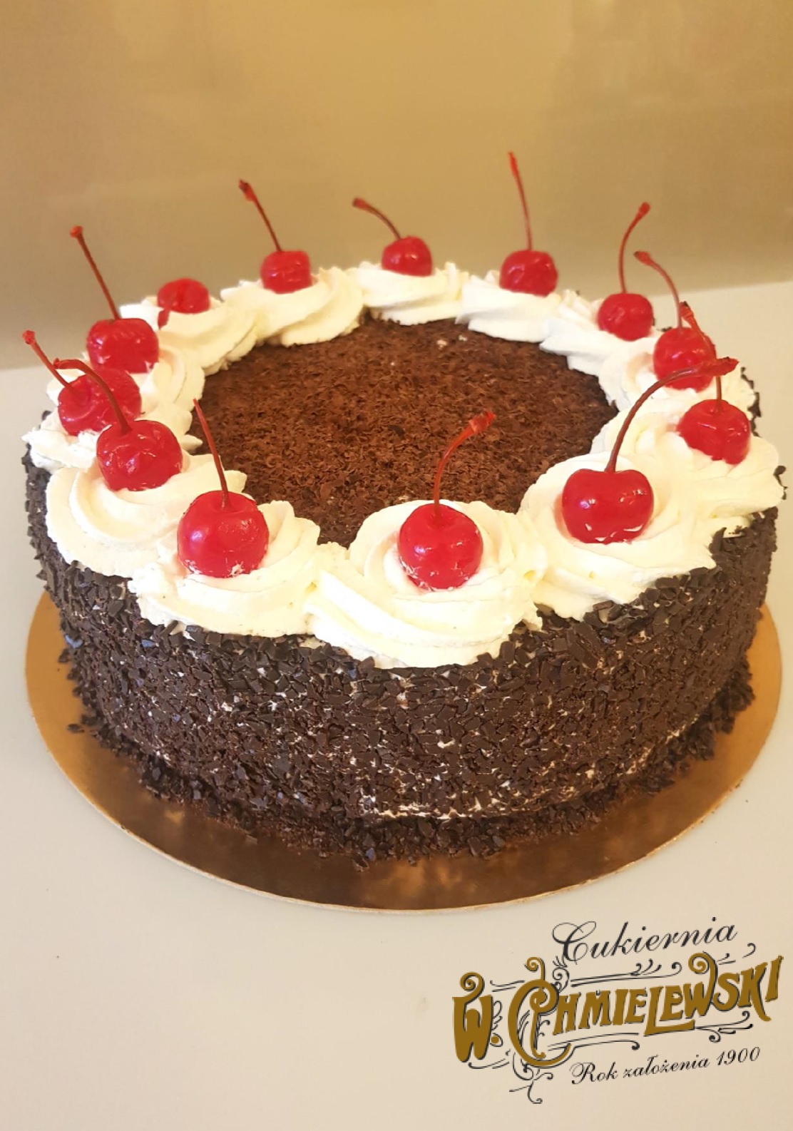 Tort Szwarcwaldzki z bitą śmietaną i wiśniami na złotym podkładzie, posypany drobnymi wiórkami czekolady, z widocznym logo cukierni w prawym dolnym rogu.