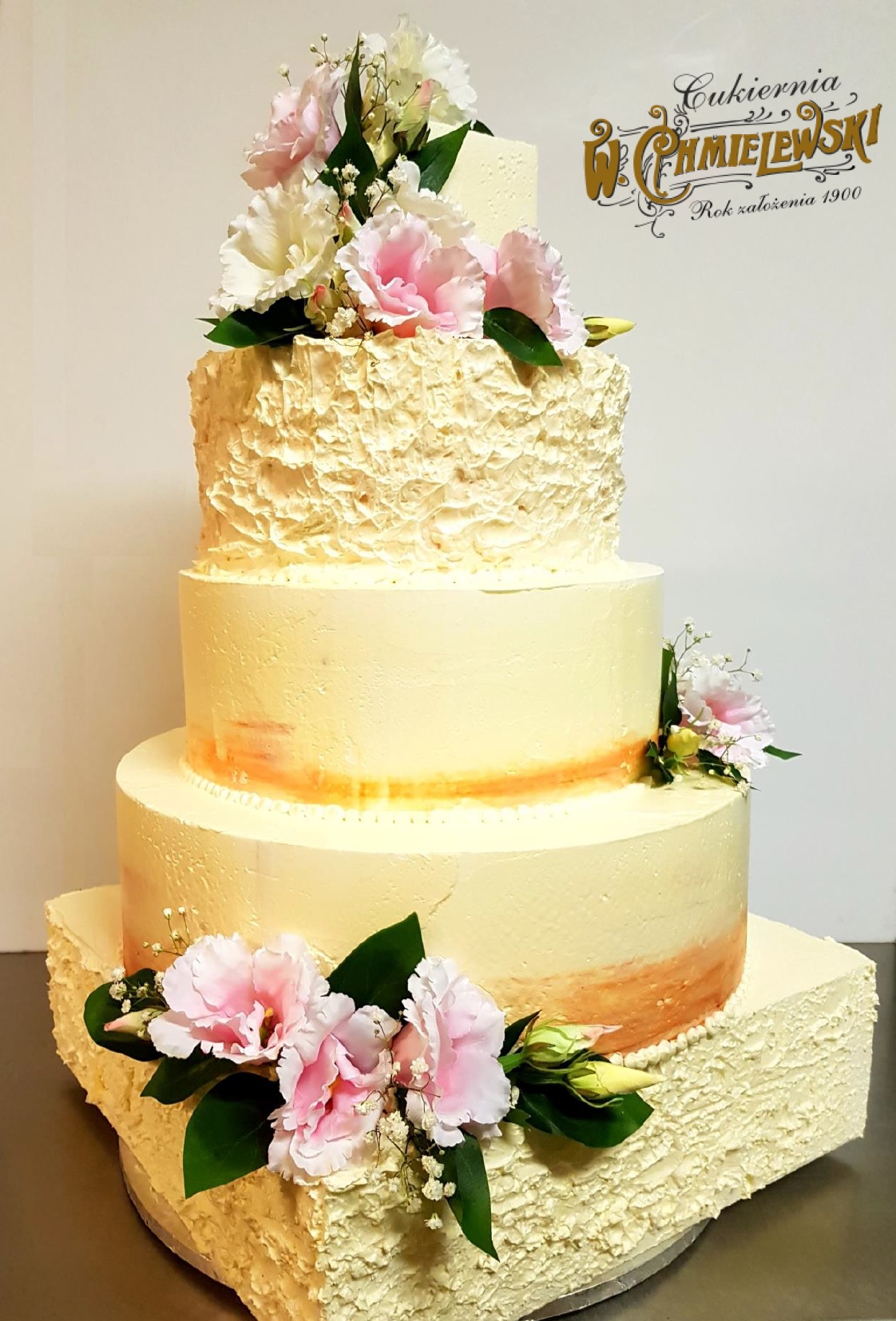 Elegancki, czteropiętrowy tort weselny w odcieniach kremu i brzoskwini, udekorowany świeżymi, różowymi i białymi kwiatami, wykonany przez cukiernię z Lublina. Na górze widoczne logo cukierni.