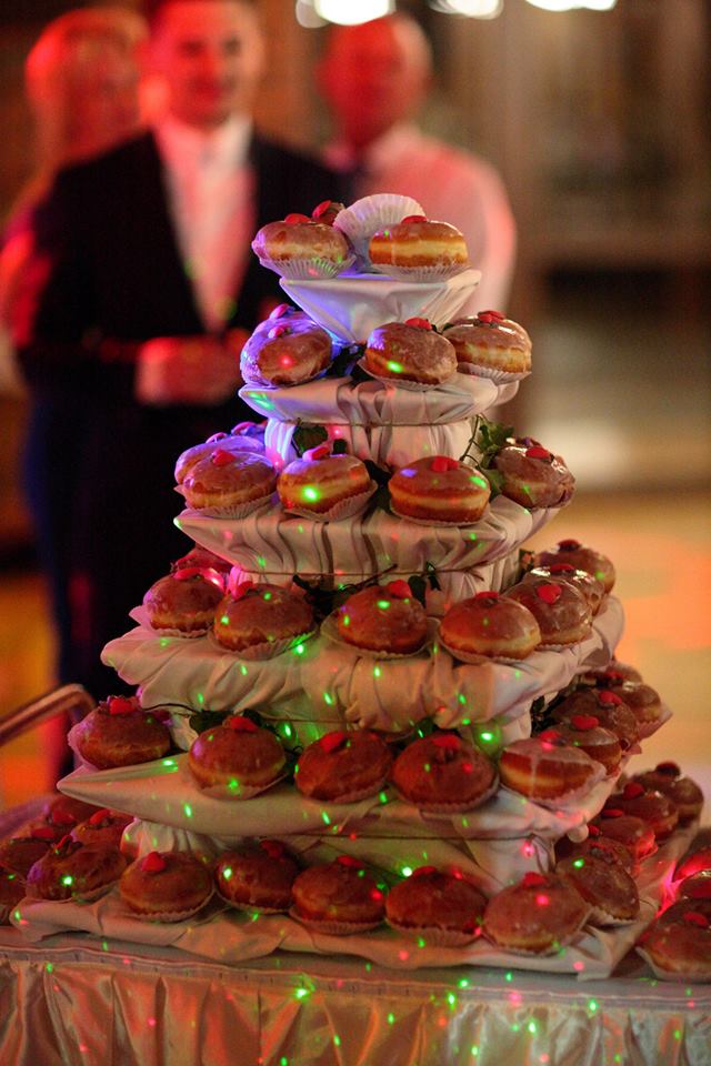 Wielopoziomowa piramida pączków z lukrem i czerwoną dekoracją, podświetlona kolorowymi światełkami, w tle osoby w strojach wieczorowych.