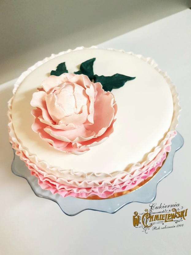 Tort zdobiony kremową różą i falbankami na szklanym paterze, z widocznym logo piekarni w rogu.