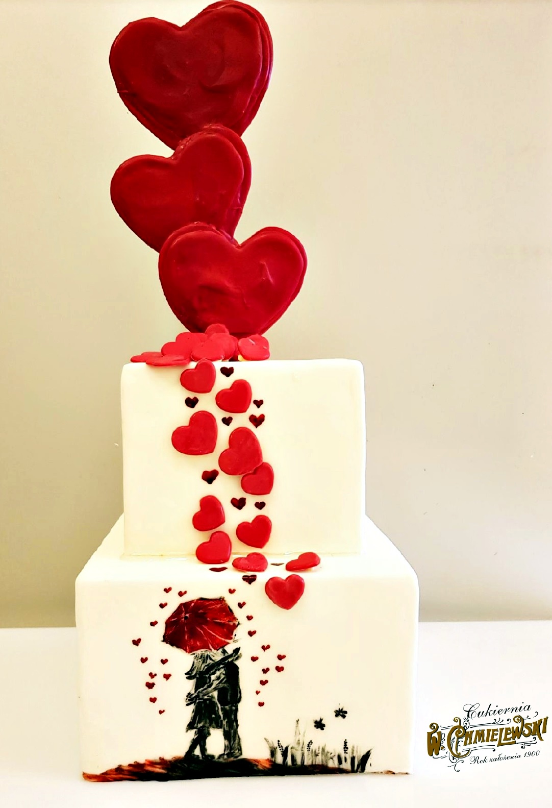 Dwupiętrowy tort kwadratowy z białym lukrem, ozdobiony czerwonymi sercami i ręcznie malowaną sceną pary pod czerwonym parasolem, z logo firmy na dole.