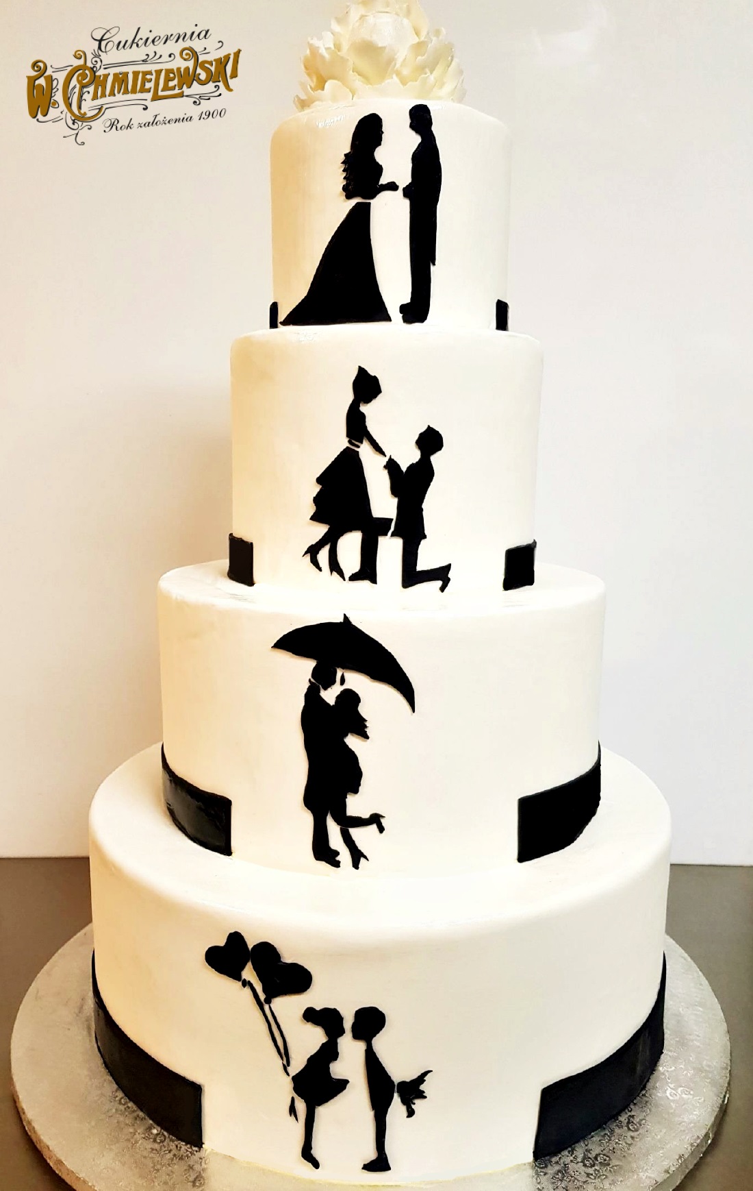 Czteropiętrowy tort weselny z białym lukrem, ozdobiony czarnymi sylwetkami pary w różnych etapach związku: ślub, zaręczyny, taniec pod parasolem i dzieci z balonami w kształcie serc. Na szczycie...