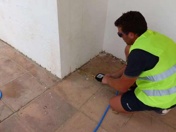 Mężczyzna w okularach i kamizelce odblaskowej, klęczący na brązowej podłodze z płytek, mierzy wilgotność ściany przy podłodze za pomocą elektronicznego miernika z sondą.