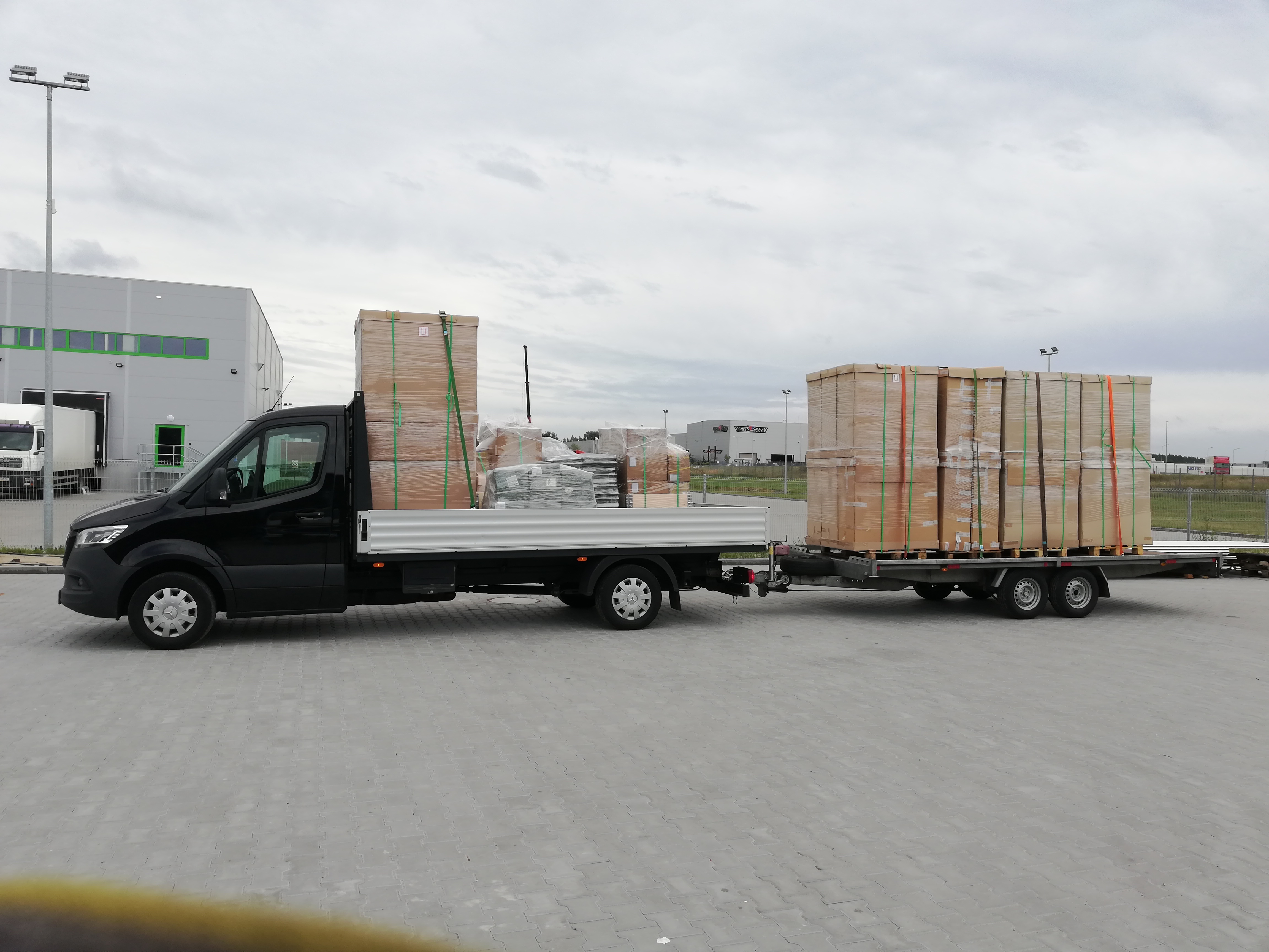 Czarny bus z platformą transportową przewozi zabezpieczone palety z towarem na betonowym placu przed budynkiem magazynowym.